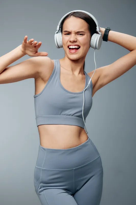 Fitness, woman in athletic wear dancing mid workout with headphones, energetic movement against a neutral backdrop. Active lifestyle and music powered training in a modern vibe.