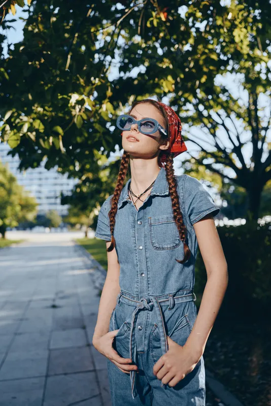 Modern boho-western denim street style woman wearing sunglasses and a red bandana, embracing a carefree lifestyle with braided hair and natural urban background in film color.