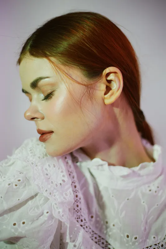 Portrait of a serene young woman with fair skin and auburn hair, dressed in a vintage white blouse against a soft pastel background, embodying natural beauty and calmness