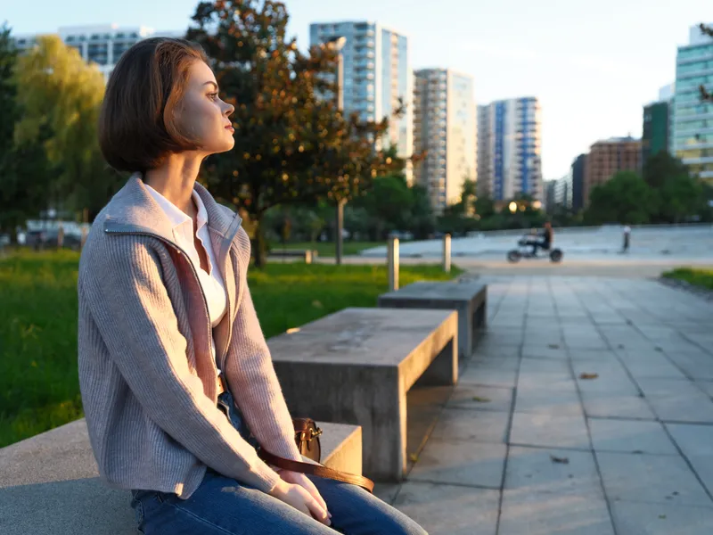 A thoughtful young woman sits on a park bench in an urban setting, enjoying a moment outdoors as modern buildings rise in the background, creating a calm, reflective mood.