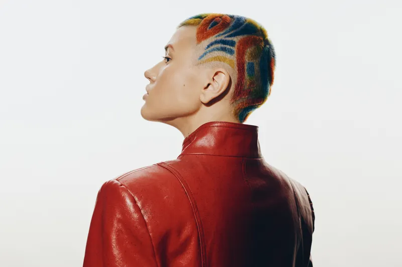 Stylish woman with colorful short hair and intricate shaved patterns wearing a vibrant red leather jacket standing confidently against a white background.