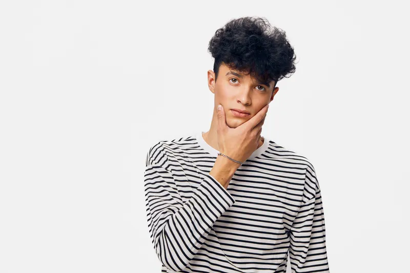 thoughtful man wearing striped shirt touching chin with hand, posing against white background with curly hair and casual style, portrait concept