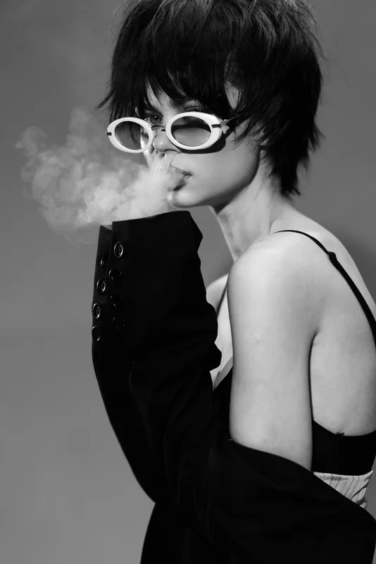 Black and white portrait of a young woman exhaling smoke wearing stylish sunglasses and a jacket sleeve over her hand.