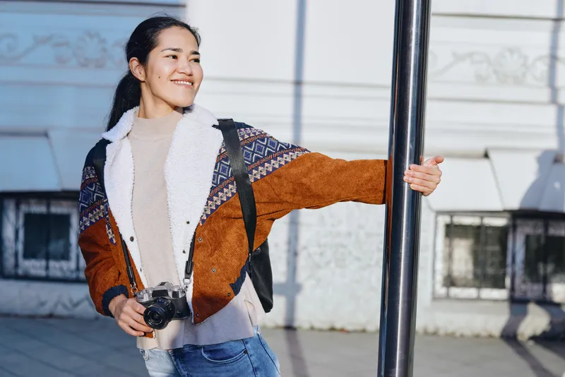 Woman with camera smiles while holding a street pole, exploring urban photography and casual streetwear on a sunny day, candid moment in motion