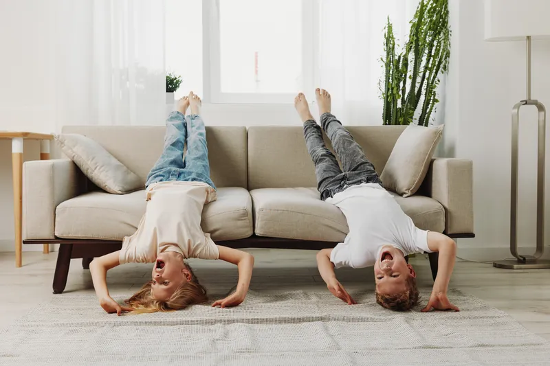 Two happy children play upside down on a beige sofa in a bright living room with natural light and indoor plants. Concept of childhood fun, home leisure, and family happiness.