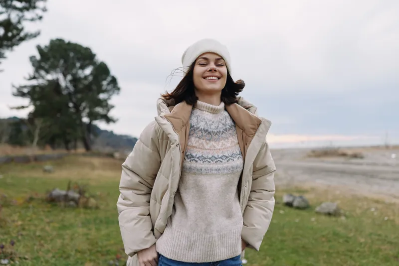 Woman outdoor portrait with a smiling face wearing sweater and jacket in a field landscape, trees and sky visible, conveying casual fashion and relaxed lifestyle