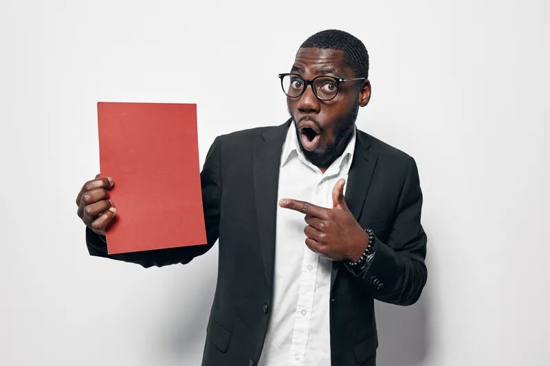 African American man with dark skin wearing glasses and black suit holding red paper and pointing with surprised expression on white background in studio.