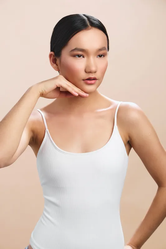 Asian woman with a calm expression poses in a studio against a soft beige backdrop, wearing a white tank top. The image conveys elegance, simplicity, and confident poise for lifestyle and fashion