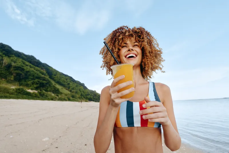 Woman with curly hair on beach by sea in summer, holding a colorful drink with straw, smiling and laughing, relaxed vacation mood, blue sky and green hills