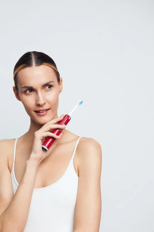 Bright studio portrait of a young woman with blonde hair wearing a white tank top, brushing teeth with a red toothpaste tube, soft smile, clean background, copy space