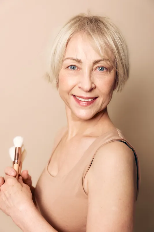 Smiling mature woman with short blonde hair holds makeup brush, wearing beige top against neutral background. Concept of beauty, skincare, self-care, natural look, and aging gracefully.
