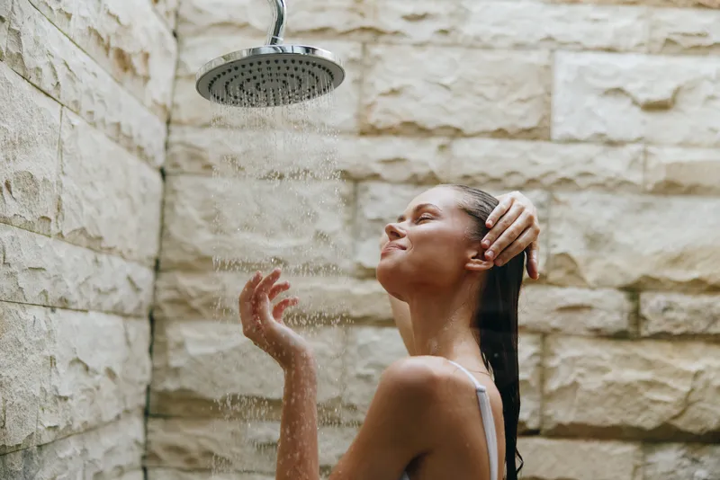 woman showering outdoors under rain showerhead with stone wall background, enjoying refreshing water and natural spa feeling in warm daylight