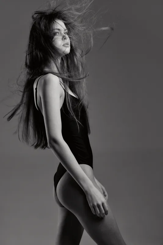 Black and white side profile of a young woman with flowing hair wearing a black bodysuit against a plain background