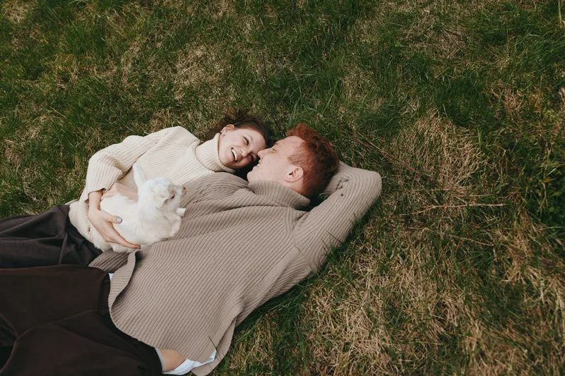 Warm couple lying outdoors on grass, embracing under soft sunlight, a cat resting beside them, cozy blanket, relaxed mood, candid moment, natural colors, peaceful countryside vibe
