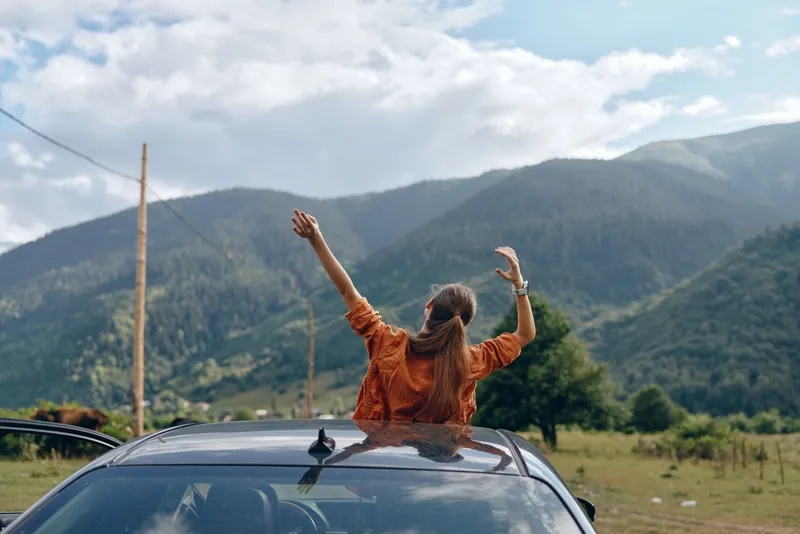 Woman stands on a car roof with arms raised enjoying freedom during a road trip in a scenic mountain landscape outdoors, conveying adventure and happiness on the open road