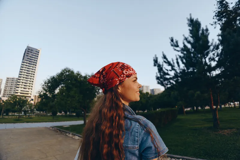 Lifestyle woman in modern boho-western denim street style wearing red bandana, enjoying urban park with film color warmth and natural outdoor setting.