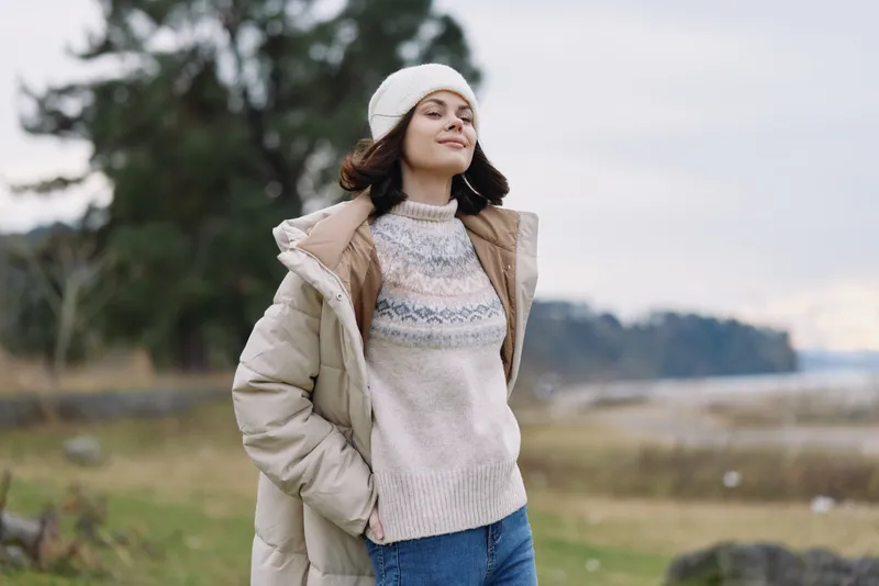 Winter outdoors, woman wearing a warm coat and knitted sweater, beanie, posing confidently in an open landscape with trees and a cloudy sky.