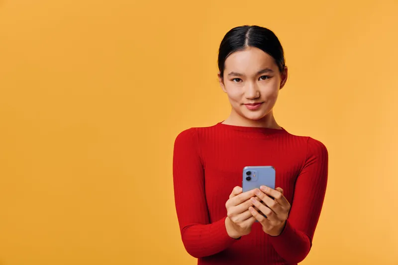 Young woman wearing red sweater holding smartphone smiling with confidence on yellow background for technology, communication and social media concept.