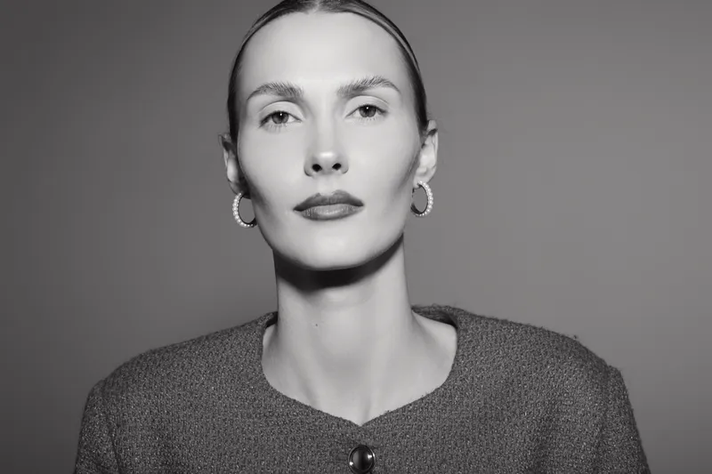 Portrait of a white woman with pale skin and slicked back hair wearing hoop earrings and a textured top against a plain grey background. Fashion and beauty concept.