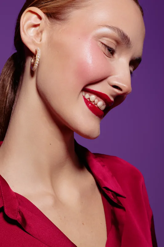 Close-up of smiling woman with red lipstick and shiny skin wearing red blouse and gold earrings on purple background. Concept of beauty, makeup, fashion, and happiness.