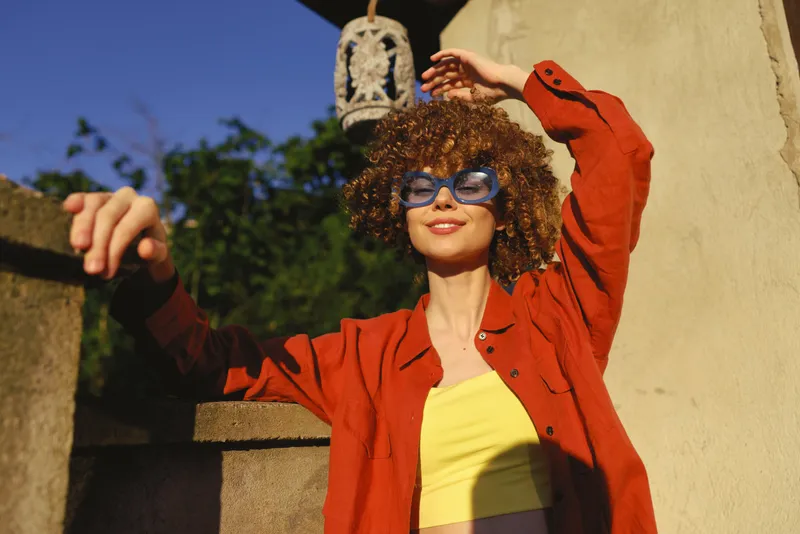 Young woman with curly hair wearing a red jacket and yellow top dances outdoors in bright sunlight