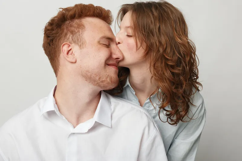 Young woman with curly hair kisses smiling man on cheek. Both wear casual shirts and show affection and happiness in a close intimate moment on a plain light background.