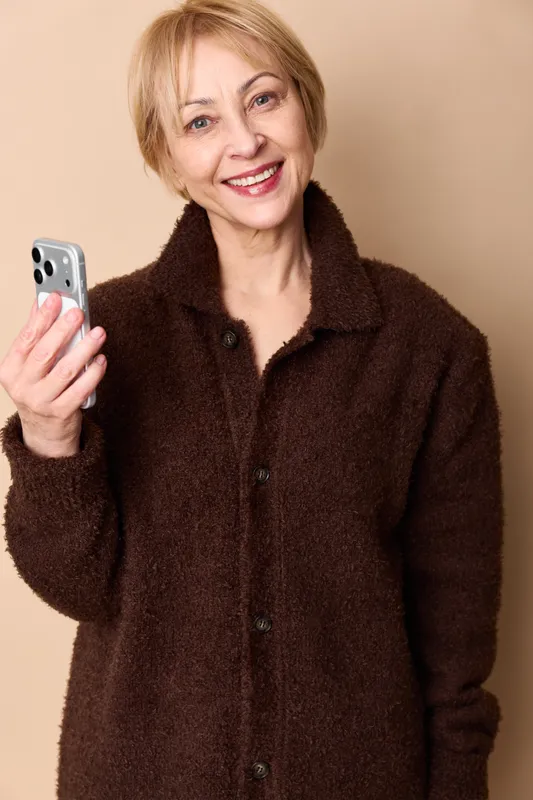 Woman, senior holding smartphone, mobile portrait, smiling, hands visible, beige background, warm indoor light, coat, friendly communication
