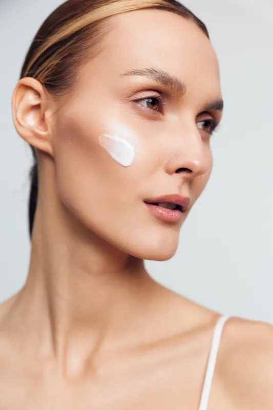 Young woman with clean skin applies white cream on cheek closeup isolated on gray background skincare beauty routine natural makeup concept.