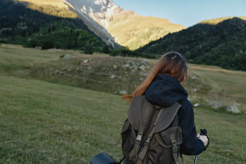 A lone hiker with a backpack stands on a grassy hillside, facing a rugged mountain range. The scene conveys adventure, solitude, and a deep connection with nature.