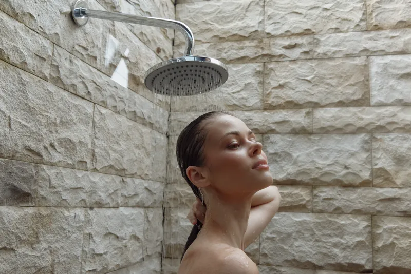 woman showering under rain showerhead with closed eyes and wet hair, natural stone wall background, relaxing and refreshing moment in bathroom
