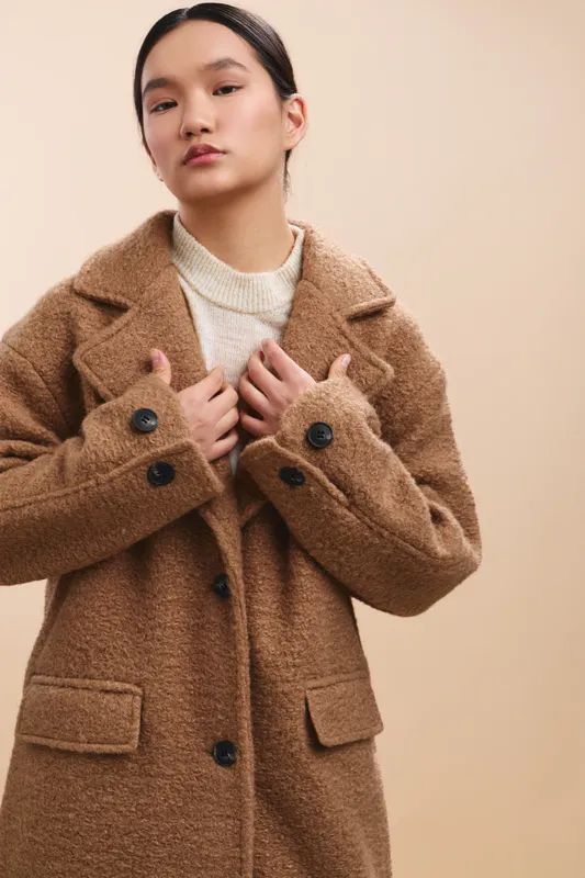 Asian woman wearing a cozy brown coat and light knit sweater stands against a neutral backdrop, offering a poised fashion portrait with calm confidence and modern style.
