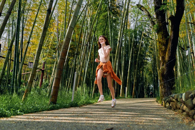 Woman running on a gravel path through a bamboo forest, smiling in casual sportswear with tied jacket, enjoying outdoor exercise, sunlight and nature trail for fitness and leisure.