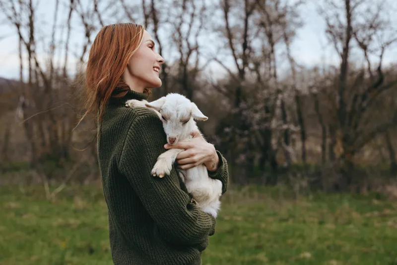 A calm outdoor scene shows a woman in a knitted sweater gently holding a young goat, with a soft rural backdrop. A moment of companionship, tenderness, and simple farm life in natural light.