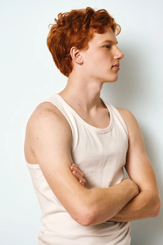 Young man with red hair and freckles wearing white sleeveless shirt standing with crossed arms looking sideways on white background. Casual confident male profile portrait in studio.