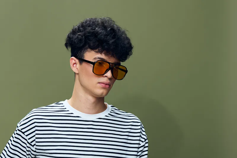 Young man wearing oversized brown sunglasses and a black-and-white striped shirt, posing confidently against a muted olive green background.