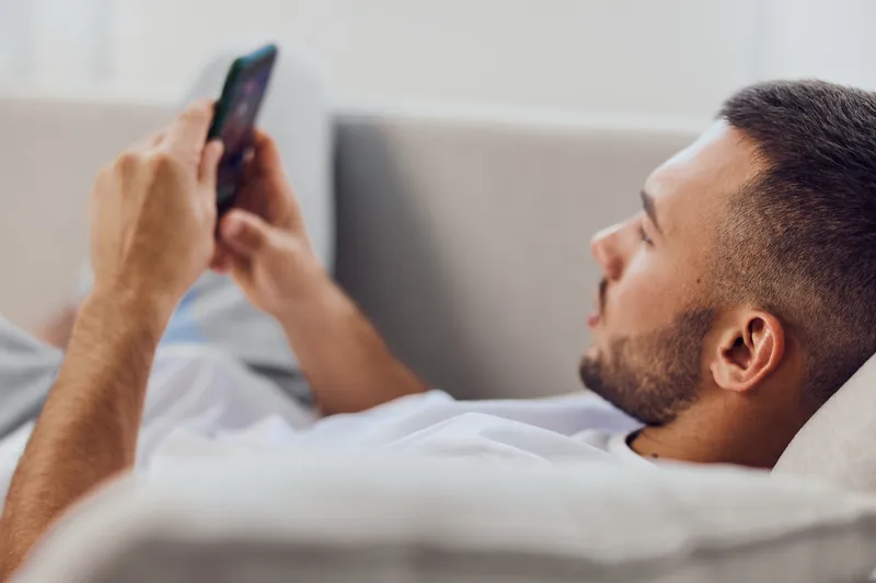 Relaxed man using smartphone while lounging on a cozy couch, dressed in casual attire. Perfect for capturing modern lifestyle moments in 2025.
