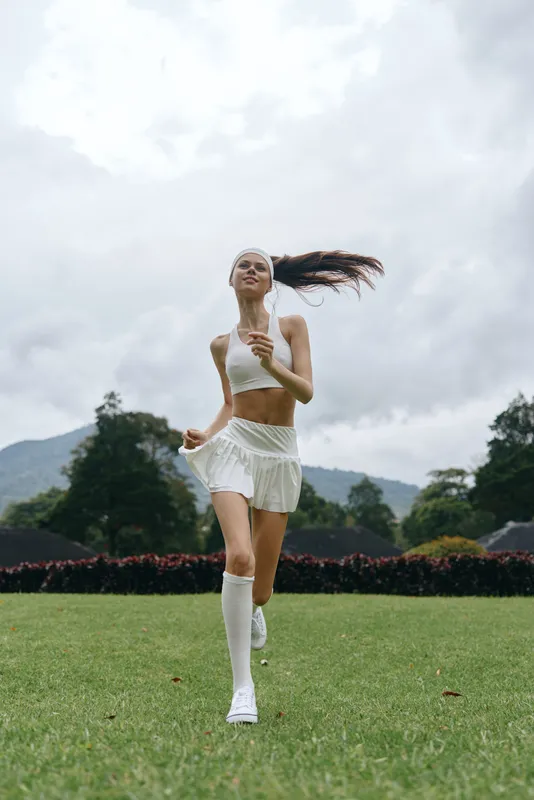 Young athletic woman in white sportswear running outdoors, expressing joy and vitality in a lush green park setting