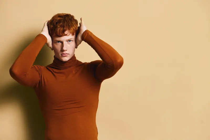 Young man with red hair and freckles wearing brown turtleneck sweater standing and posing against beige background with hands on head in studio portrait.