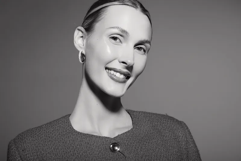 Portrait of a smiling young white woman with light skin and slicked-back hair wearing a textured top with a button. Studio lighting highlights her natural beauty and confident expression.