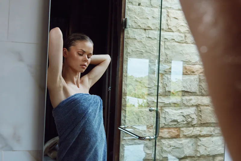 young woman wrapped in blue towel standing by a glass shower door in a modern bathroom with stone wall, morning routine, natural light, wellness and relaxation
