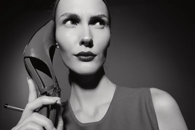 White woman with light skin holds high heel shoe near ear, posing in black and white studio portrait. Concept of fashion, communication, and creative expression.