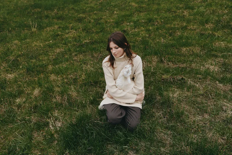 A thoughtful woman kneels on grass in a soft sweater, a small goat kid nearby. A calm pastoral moment capturing rural lifestyle, companionship with animals, and gentle moments of reflection.