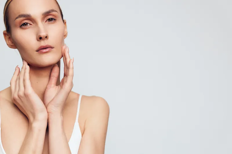 A young woman with flawless skin gently touches her neck and face, wearing a white tank top against a plain light background. Concept for skincare, beauty, and wellness.