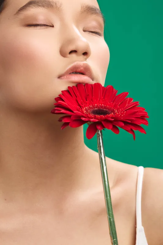Asian woman with eyes closed, gently holding a red flower near her lips in a studio setting, conveying serene beauty, poetic mood, and delicate artistic expression.