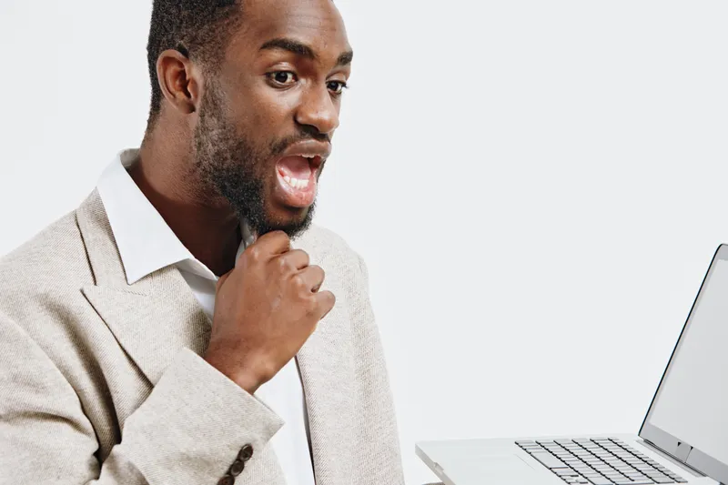 Confident young African American man in business attire looking surprised while using a laptop computer in a modern office setting. Studio shot with neutral background. People success concept