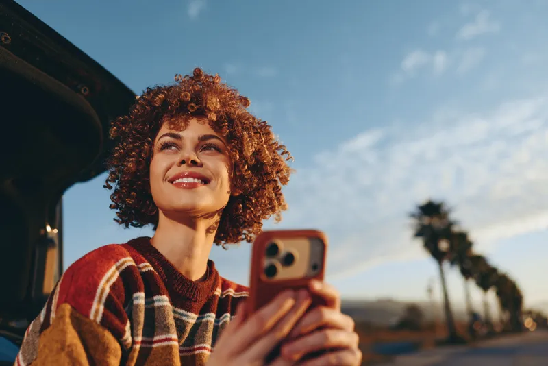 Woman with curly hair smiling while holding a smartphone inside a car at sunset, outdoors on a road, warm sunlight sky and landscape, lifestyle travel portrait