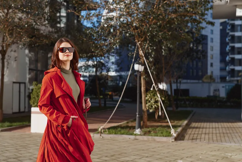 Stylish woman in a red coat walks outdoors in an urban plaza, wearing sunglasses and a confident pose, sunlit moment with light breeze and modern backdrop