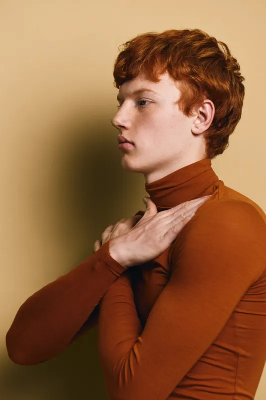Young man with red hair wearing brown turtleneck posing sideways with arms crossed on chest against beige background. Profile portrait with thoughtful expression and smooth skin tone.