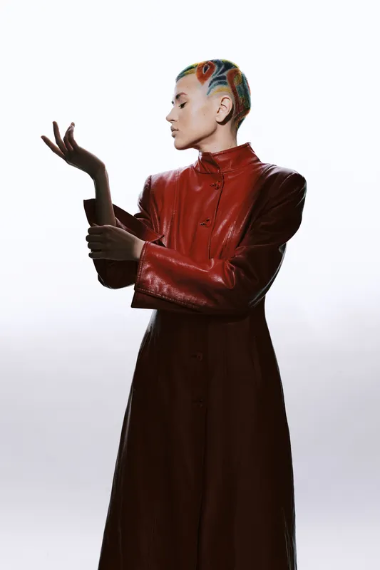 Artistic woman with colorful short hair wearing a long red leather coat posing elegantly against a bright studio background. Unique fashion and creative style expression.