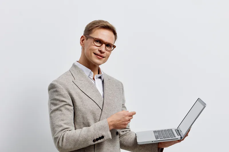 Professional smiling man with glasses in beige blazer holding open silver laptop, standing against plain light background, lifestyle concept, office work, technology, communication, modern style, posi